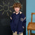 A young boy with curly brown hair stands wearing a navy blue varsity-style jacket over a matching navy polo shirt and khaki pants. The jacket features beige ribbed trim, a white embroidered laurel wreath and letter 'S' on the left chest, and striped cuffs and hem.