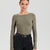 A woman modeling a long-sleeve crew-neck knit top in olive green, paired with black jeans and a black belt, against a light gray studio background.