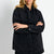 A blonde woman wearing an oversized black denim shirt jacket with a button-down front and chest pockets, posed with arms crossed against a light gray studio background.