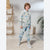 A child wearing a matching light blue sweatshirt and jogger pants set with a dinosaur print, styled with a white collared shirt underneath and white sneakers, standing in a bright, modern room.