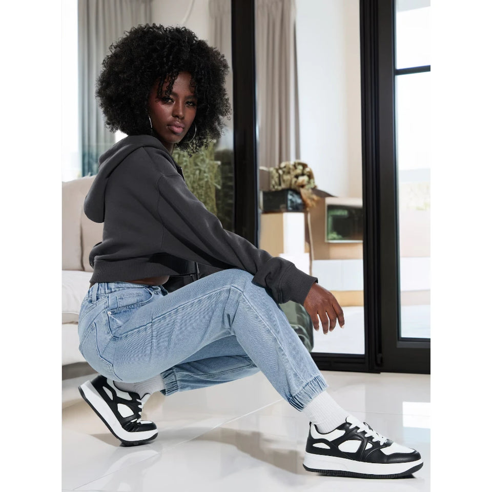 A person crouching indoors wearing a dark gray hoodie, light blue jeans, white socks, and black-and-white platform sneakers.