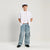A young boy modeling a casual outfit consisting of a white short-sleeve button-up shirt, light-wash wide-leg denim cargo pants, a white t-shirt, black shoes, and a white and black baseball cap, standing against a plain light gray studio background.