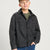A boy modeling a dark charcoal gray button-up shirt jacket over an olive green t-shirt and blue jeans, photographed against a light gray studio background.