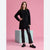 A young woman stands in a studio wearing a black ribbed long-sleeve tunic sweater, black flared pants, and black loafers, posed in front of a pink backdrop with a light blue-gray pedestal.