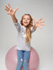Child standing on a pink exercise ball with a gray shirt and blue jeans against a plain background