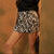 A person wearing black and beige zebra-print shorts with a black top, standing against a textured warm brown wall and holding a black clutch.