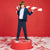 A child stands on a red pedestal against a red backdrop, wearing a navy blazer, white shirt, navy trousers, and dark sneakers, while holding a large red-and-white candy cane. White faux-snow covers the floor.