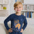 A young blond boy wearing a navy blue long-sleeve graphic shirt with a game controller design, standing in a brightly lit room with bookshelves in the background.