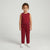 A young child stands against a light gray studio background wearing a sleeveless red top and matching red jogger-style pants with white sneakers.