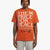 A man wearing a bright orange t-shirt with 'THE PRICE FOR PEACE' printed in large white letters, featuring a butterfly graphic and text on the front, paired with light gray pants, against a plain light gray background.