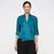 A woman modeling a teal textured wrap-style blouse with three-quarter sleeves and a decorative brooch at the waist, paired with black pleated pants, against a plain light gray background.