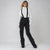 A woman modeling black wide-leg ski pants with black suspenders, paired with a white turtleneck sweater and black sneakers, against a light gray studio background.