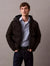 A man wearing a black hooded puffer jacket over a light blue button-down shirt and dark jeans, standing against a plain gray studio background.