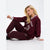 A woman sitting on a light gray studio floor wearing a matching burgundy sweatshirt and jogger pants set with white varsity-style trim and lettering.