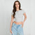 A woman modeling a light gray short-sleeve crop top with a split neckline and button detail, paired with light blue jeans, against a plain light gray studio background.
