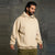 A man wearing a light beige pullover hoodie with a kangaroo pocket and subtle chest lettering, paired with olive green pants, standing against a textured gray concrete wall.