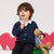 A young child with brown hair sits on a green block wearing a navy blue cable-knit cardigan over a white collared shirt, paired with black pants. The child is holding a brown teddy bear and is posed against a white background with pink heart-shaped props.