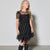 A young girl with blonde hair wearing a black tiered tulle dress with sheer mesh sleeves and black knee-high boots, standing against a light gray studio wall.