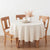 An off-white round tablecloth draped over a wooden dining table with three matching wooden chairs, styled with a white vase of dried grass and plates on a light wood floor against a white wall.