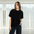 A woman wearing a plain black short-sleeve crewneck t-shirt and dark blue jeans, standing in a bright room with sheer white curtains and a blurred mountain background.