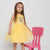 A young girl with glasses and a pink headband stands next to a pink chair wearing a yellow sleeveless dress with a leopard-print heart graphic and the words 'world heart'.