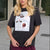 A blonde woman wearing a dark charcoal graphic t-shirt with a white square print featuring cosmetics and a coffee cup, paired with burgundy pants, standing outdoors on a paved walkway.