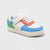 A toddler's white low-top sneaker with colorful accents in light blue, coral red, green, and yellow, featuring a hook-and-loop strap closure and white laces, shown on a light gray background.