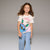 A young girl with long brown hair stands against a neutral gray studio background wearing a white graphic t-shirt with a blue cartoon character and the text 'Stitch - may DOO!', light blue wide-leg jeans, and pink sneakers.