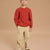A young boy wearing a rust-red cable-knit sweater and beige corduroy pants, standing against a solid warm beige studio background.