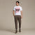 A man modeling a white crewneck t-shirt with a red and black graphic print that reads 'LOOK OUT' with a camera illustration, paired with brown jogger pants and brown sneakers, against a solid beige studio background.