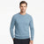 A man wearing a light blue crewneck knit sweater and dark trousers, standing against a plain white studio background.