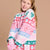 A young girl with blonde hair smiling while wearing a pink and white holiday-themed knit sweater with green and blue accents, set against a solid beige background.