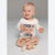 A smiling toddler sitting cross-legged on a light gray studio background, wearing a white long-sleeve shirt with the words 'COOKIE BAKING CREW' in black and orange lettering, paired with beige pants printed with gingerbread cookies and candy canes.