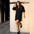 A woman wearing a black puff-sleeve button-front mini dress with a white headband, posing against a beige stone wall.