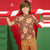 A young boy wearing a brown short-sleeve t-shirt with a gingerbread man and candy cane pattern, posing with a giant candy cane prop in front of a festive red and green holiday backdrop.