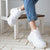 White chunky platform sneakers worn by a person sitting on a white wooden stool, paired with blue jeans.