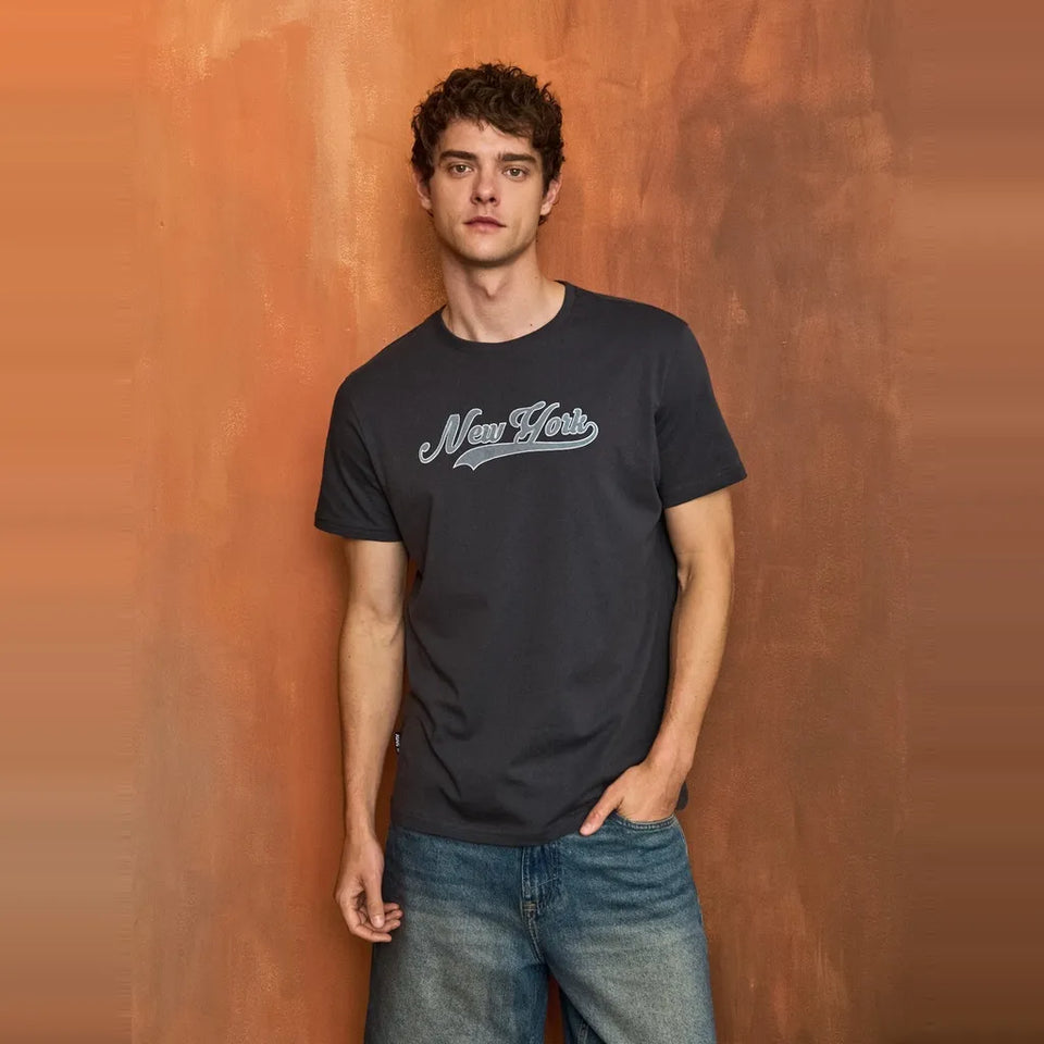 A man wearing a dark charcoal gray t-shirt with a white 'New Heirs' logo across the chest, paired with blue jeans, standing against a textured orange-brown wall.