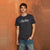 A man wearing a dark charcoal gray t-shirt with a white 'New Heirs' logo across the chest, paired with blue jeans, standing against a textured orange-brown wall.