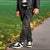 A person outdoors wearing dark gray washed denim jeans, a black leather jacket, a black and white striped shirt, and black and white sneakers, standing on a paved path next to a grassy park area.