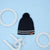A dark navy blue cable-knit beanie with a matching pom-pom, featuring two horizontal white stripes near the brim and a small brown leather brand tag. The beanie is displayed on a light blue background with white geometric props.