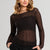 A woman wearing a dark brown sheer lace long-sleeve top with a boat neckline, paired with black pants, against a solid light gray background.