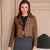 A woman modeling a cropped brown faux-suede moto jacket with silver-tone hardware, worn over a black crop top and a gray skirt, set against a beige architectural background.