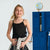 A young girl with blonde hair stands smiling in a black camisole, dark cargo pants, and a light gray sweatshirt tied around her waist, posed in front of blue lockers with a globe on top.