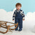 A toddler stands in a snowy studio setting wearing a navy blue puffer snowsuit over a navy and cream patterned long-sleeve top, with blue boots. A wooden sled is to the left, and the background features a light blue sky and white cloud cutouts.