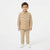 A young boy modeling a beige two-piece outfit consisting of a long-sleeve button-up shirt and matching trousers, paired with white sneakers, against a plain white studio background.