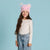 A young girl with long brown hair smiling, wearing a pink unicorn-themed beanie with colorful fluffy ears, a cream long-sleeve top with a small heart pattern, and light blue jeans, set against a solid light blue background.