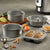 A set of stainless steel baking pans and a loaf pan shown on a gray countertop with baking ingredients and a partially baked double loaf inside. The set includes two round springform pans, a bundt pan, and a rectangular loaf pan with baked bread.