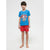 A young boy stands barefoot against a light gray studio background, wearing a bright blue Superman graphic T-shirt and red shorts with a Superman logo patch.