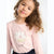 A young girl with curly brown hair wearing a light pink long-sleeve t-shirt with a white and yellow flower applique on the front, paired with dark navy pants, set against a plain white background.