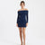 A woman modeling a navy blue off-the-shoulder long-sleeve mini dress against a plain white studio background.